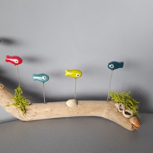 Könnte beinhalten: Ein Treibholzast mit fünf Keramikfisch-Ornamenten in verschiedenen Farben, darunter Rot, Blau, Gelb und Grau. Die Fische haben weiße Augen und sind mit Draht am Ast befestigt.