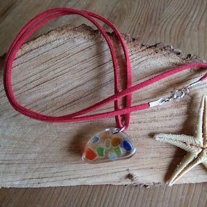 Puede incluir: Un collar de cordón de gamuza rojo con un cierre plateado y un colgante de vidrio transparente lleno de piezas de vidrio de mar de colores.