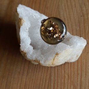 Puede incluir: Un anillo de oro con un engaste de resina transparente que contiene escamas doradas. El anillo está sobre un cristal blanco.