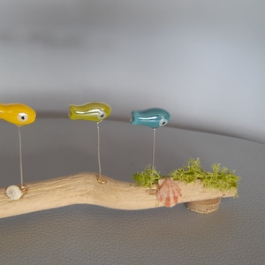 Puede incluir: Tres pequeñas figuritas de peces de cerámica en amarillo, verde y azul están montadas en una rama de madera flotante con musgo y conchas.