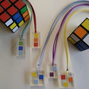 Peut inclure: Cinq colliers colorés avec des pendentifs carrés. Chaque pendentif a une combinaison de couleurs différente de bleu, rouge, jaune, vert et orange. Les colliers sont faits de cordons de différentes couleurs.