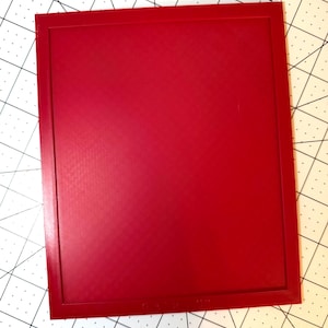 May include: A rectangular red plastic frame with a textured surface, positioned on a white cutting mat with black grid lines and a Westcott ruler visible at the top, showing a width of approximately 2.5 inches for the frame's top edge.