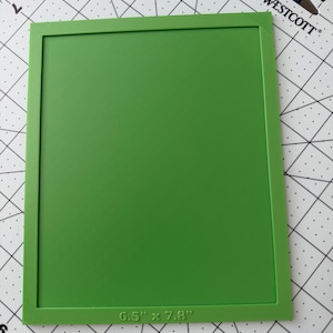 May include: A rectangular green frame with a flat surface, featuring the text 6.5" x 7.8" embossed on the bottom border. The frame is positioned on a white cutting mat with black grid lines and angle markings, showing a Westcott brand logo.