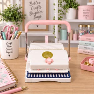 May include: A white and pink binding machine with a large handle, featuring a central Heidi Swapp logo, a pink adjustment knob, and a dark blue base with white vertical slots, designed for punching holes in paper for spiral-bound projects.