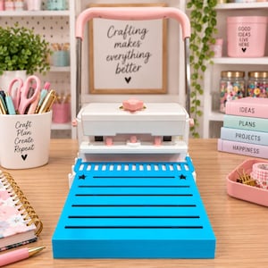 Peut inclure: Une perforatrice à papier blanche et rose pâle avec une base bleu vif dotée de fentes horizontales et d'étoiles. L'appareil possède une poignée en métal et un bouton rose central, conçu pour les travaux manuels sur un bureau.