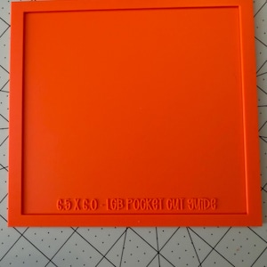 May include: A bright orange square cutting guide with a raised border. The text 6.5 X 6.0 - LGB POCKET CUT GUIDE is embossed along the bottom edge of the frame. This tool is designed for precise material alignment and cutting tasks.