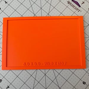 May include: A rectangular orange frame with a flat surface, featuring the embossed text 8.5 X 5.5 - 215.9 X 139.7 at the bottom. The object is positioned on a cutting mat with grid lines, showing a clear, solid orange color and a smooth finish.
