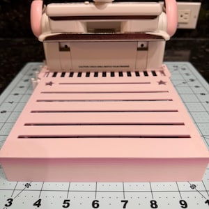 May include: A light pink paper punch tool with a white top section and metallic handle accents. The base features horizontal slots and star cutouts, positioned on a cutting mat with visible inch markings ranging from 3 to 9 inches in width.