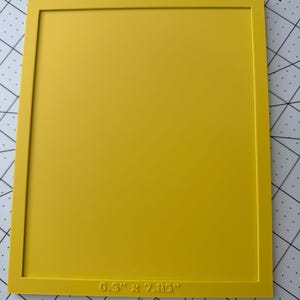 May include: A rectangular yellow frame with a smooth, flat surface. The bottom edge of the frame features embossed text that reads 6.5" x 7.85", indicating the dimensions of the inner opening. The frame is positioned on a white cutting mat.