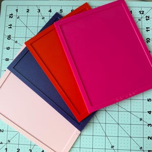 May include: A set of four rectangular silicone frames in pink, dark blue, red, and magenta, each featuring a raised border. The magenta frame is clearly marked with the dimensions 6.5 inches by 8.0 inches, providing a clear size reference.