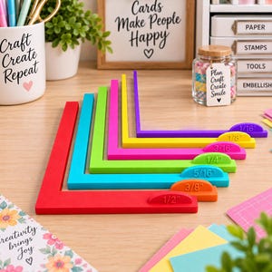 May include: A set of seven L-shaped plastic paper crafting alignment tools in red, orange, turquoise, lime green, pink, yellow, and purple. Each tool features a raised tab with a printed measurement ranging from 1/16 inch to 1/2 inch.