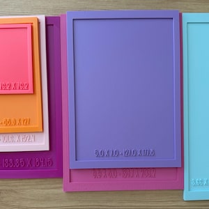 Peut inclure: Une collection de moules rectangulaires en silicone de différentes couleurs, notamment violet, rose, orange et bleu clair. Chaque moule présente un texte en relief indiquant les dimensions en pouces et millimètres, comme 3.0 x 3.0 - 76.2 x 76.2.