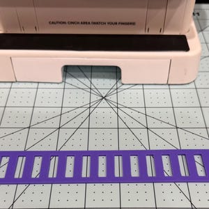 May include: A light pink binding machine with a black strip featuring the text CAUTION: CINCH AREA (WATCH YOUR FINGERS) and a separate flat purple plastic guide with vertical slots and two small mounting holes at the ends.