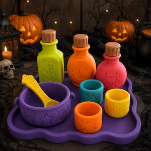 May include: A set of silicone potion-making toys including a purple tray, a purple bowl with a yellow spoon, three small cups in teal, orange, and yellow, and three bottles in lime green, orange, and pink, all featuring embossed symbols.
