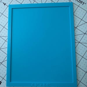 May include: A rectangular bright blue silicone mat with a raised border frame. The bottom edge of the mat features the embossed text 6.5" x 8.0", indicating the dimensions of the product. The mat has a smooth, flat surface and uniform color.