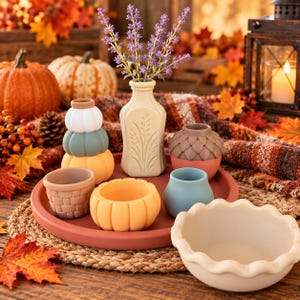 May include: A collection of small, colorful silicone containers shaped like pumpkins, acorns, and a basket, arranged on a round terracotta tray with a cream-colored vase holding lavender sprigs and a separate scalloped-edge bowl.