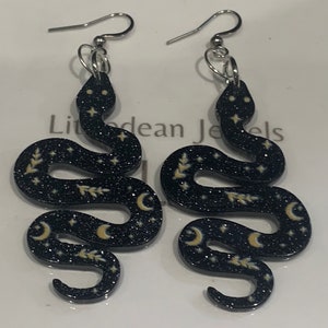 May include: A pair of black snake earrings with gold glitter and moon and star accents. The earrings are made of acrylic and have silver hooks.