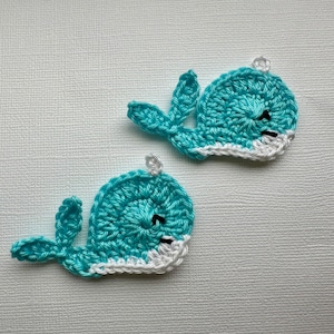 May include: Two crocheted whale appliques in turquoise blue with white trim. The whales have black eyes and a simple smiling mouth.
