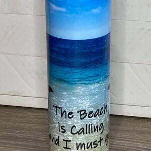 May include: A white stainless steel tumbler with a blue ocean and beach scene. The text "The Beach is Calling and I must go" is printed on the tumbler.