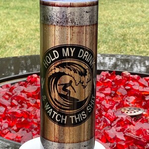 May include: A stainless steel tumbler with a wood grain design and a black circle with the text "Hold My Drink & Watch This Shit" and a silhouette of a surfer riding a wave.