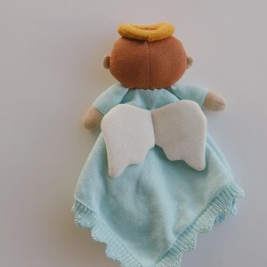 May include: A knitted baby angel doll with a light blue blanket. The doll has a brown head with a yellow halo, beige arms, and white wings. The blanket has a scalloped edge.