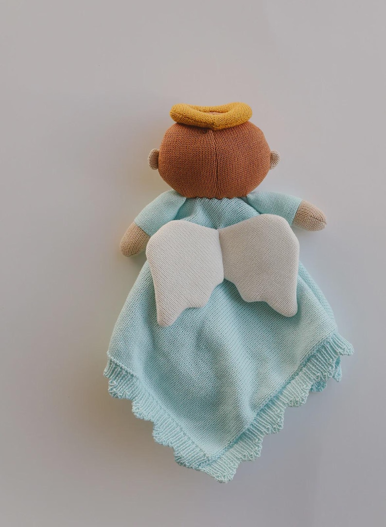 May include: A knitted baby angel doll with a light blue blanket. The doll has a brown head with a yellow halo, beige arms, and white wings. The blanket has a scalloped edge.