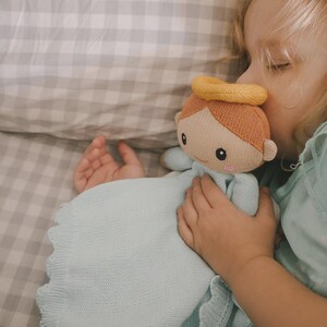 May include: A soft, knitted angel doll with a yellow halo and light brown hair. The doll is holding a light blue blanket with a scalloped edge. The doll is being held by a child.