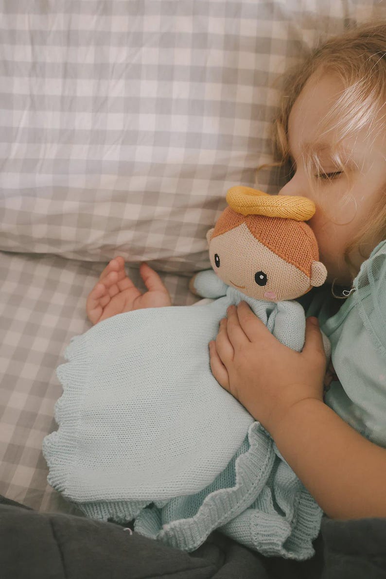 May include: A soft, knitted angel doll with a yellow halo and light brown hair. The doll is holding a light blue blanket with a scalloped edge. The doll is being held by a child.