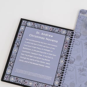 May include: A spiral-bound notebook with a light purple cover and pages. The open page displays text: "St. Andrew Christmas Novena." The border features a floral design in shades of purple and white. The adjacent page has a repeating pattern of line drawings.