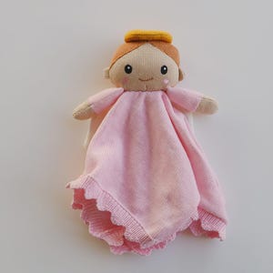 May include: A knitted angel doll with a light pink dress and a yellow halo. The doll has a smiling face, brown hair, and small arms. The dress has a scalloped edge.