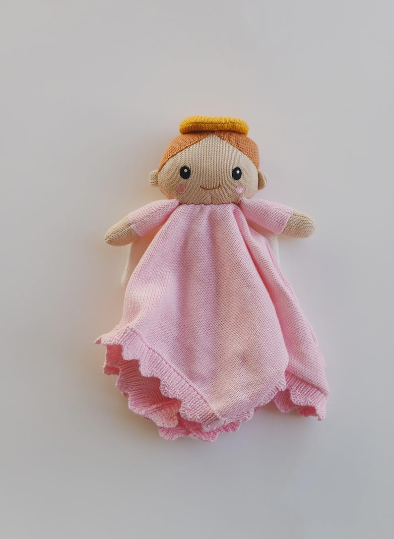 May include: A knitted angel doll with a light pink dress and a yellow halo. The doll has a smiling face, brown hair, and small arms. The dress has a scalloped edge.