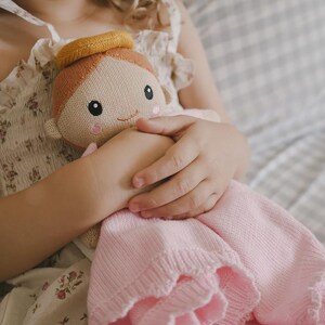 May include: A knitted doll with a smiling face, wearing a yellow halo and a pink dress. The doll is held in a child's hands. The doll's hair is light brown, and the dress has a ruffled edge. The background is a soft, neutral color.