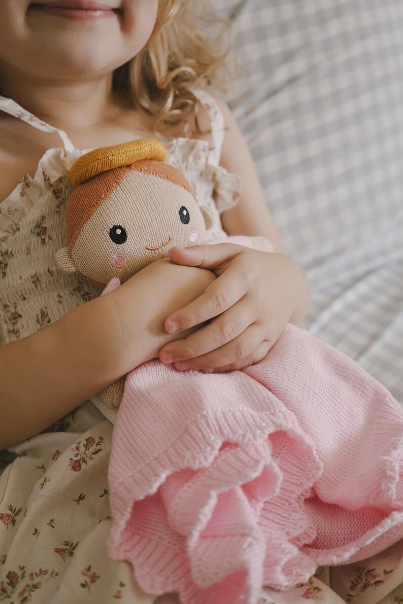 May include: A knitted doll with a smiling face, wearing a yellow halo and a pink dress. The doll is held in a child's hands. The doll's hair is light brown, and the dress has a ruffled edge. The background is a soft, neutral color.