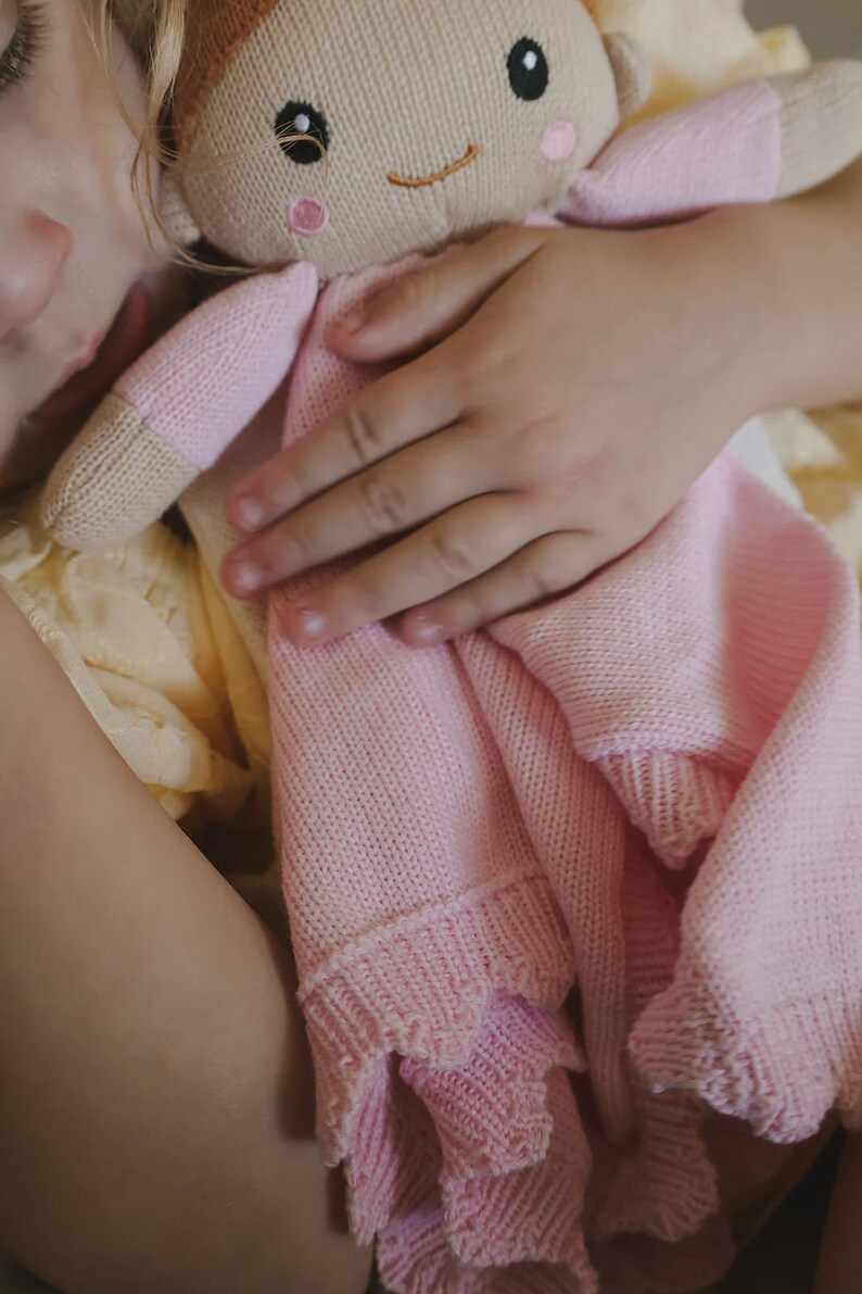 May include: A soft, knitted doll with a smiling face, rosy cheeks, and brown hair is held in a child's arms. The doll wears a pink dress and has matching pink arms. The child is wearing a yellow top.