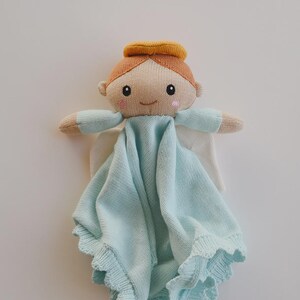 May include: A knitted angel-shaped baby comforter in soft pastel colors. The doll has a tan face, orange hair, and a yellow halo. The angel wears a light blue blanket with a scalloped edge and white wings.