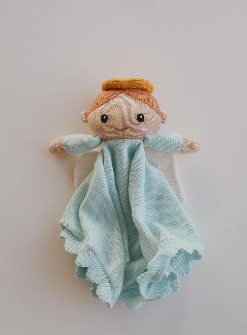 May include: A knitted angel-shaped baby comforter in soft pastel colors. The doll has a tan face, orange hair, and a yellow halo. The angel wears a light blue blanket with a scalloped edge and white wings.
