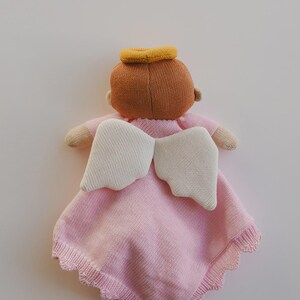 May include: A soft, knitted angel doll in pink with cream-colored wings and a yellow halo. The doll has a brown head and small arms. The pink blanket has a scalloped edge. A comforting and decorative item.