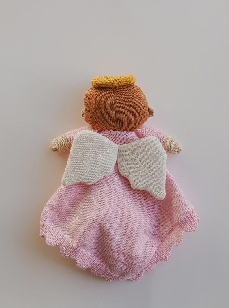 May include: A soft, knitted angel doll in pink with cream-colored wings and a yellow halo. The doll has a brown head and small arms. The pink blanket has a scalloped edge. A comforting and decorative item.