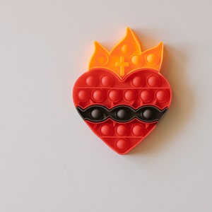 May include: A red heart-shaped pop-it toy with a black and white design and a yellow flame on top. The toy has a cross in the center of the flame.