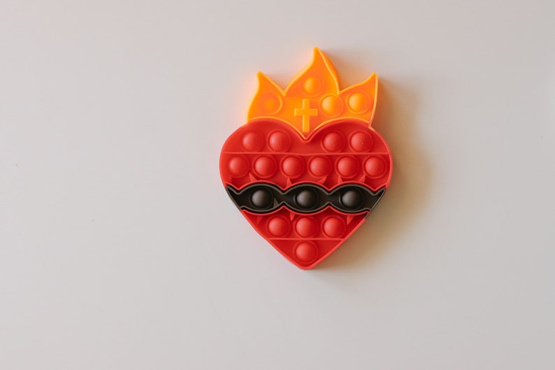 May include: A red heart-shaped pop-it toy with a black and white design and a yellow flame on top. The toy has a cross in the center of the flame.