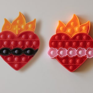 May include: Two red heart-shaped pop-it toys with a flame design on top. One has a black band around the middle and the other has a pink band.