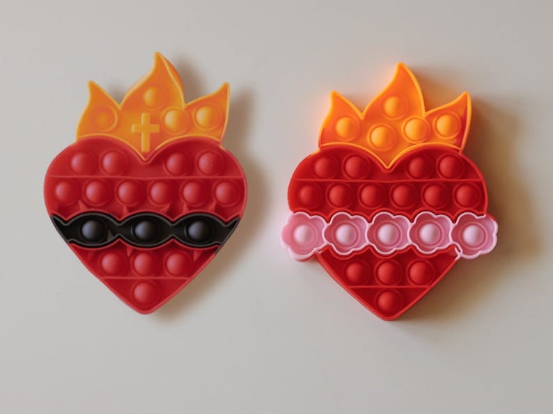 May include: Two red heart-shaped pop-it toys with a flame design on top. One has a black band around the middle and the other has a pink band.
