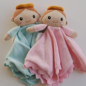 May include: Two knitted baby dolls, each with a halo, smiling faces, and soft blankets. One doll has a light blue blanket, and the other has a pink blanket. The dolls are designed with a simple, charming aesthetic.