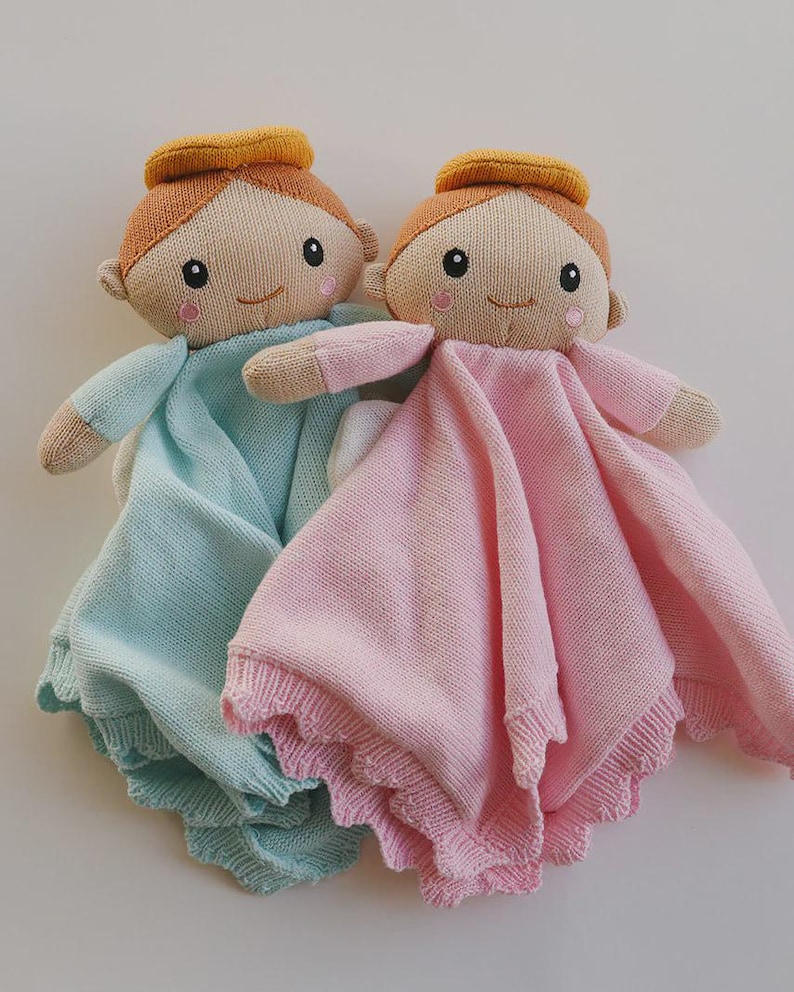 May include: Two knitted baby dolls, each with a halo, smiling faces, and soft blankets. One doll has a light blue blanket, and the other has a pink blanket. The dolls are designed with a simple, charming aesthetic.