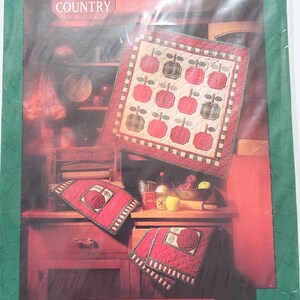 May include: A package featuring a quilted apple-themed design. The design includes a small quilt and two matching pot holders with red and green apples. The text "APPLE COUNTRY" and "MUMM'S THE WORD" are visible.