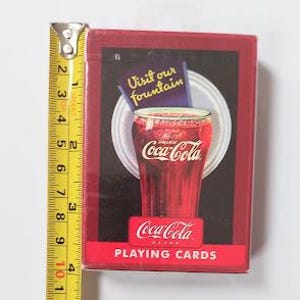 K&ouml;nnte beinhalten: Eine rote Coca-Cola Spielkartenbox mit einem Vintage-Bild eines Coca-Cola-Glases. Die Box ist etwa 9 cm hoch, mit einem gelben und silbernen Ma&szlig;band daneben.