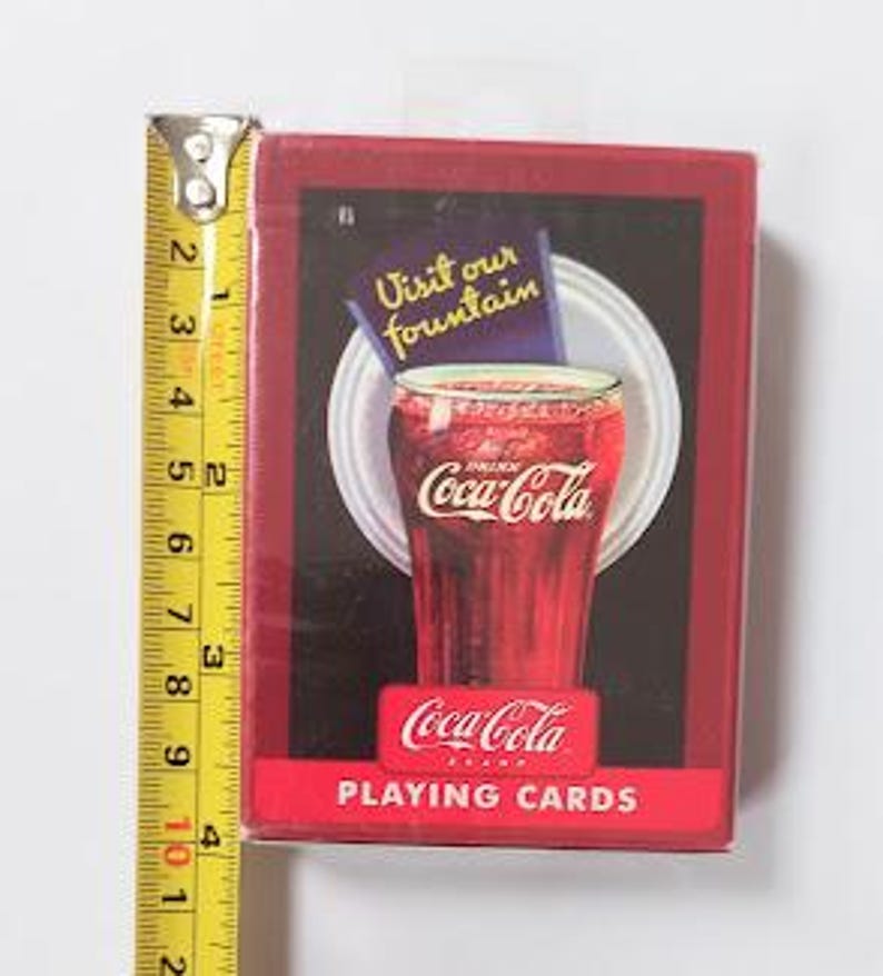K&ouml;nnte beinhalten: Eine rote Coca-Cola Spielkartenbox mit einem Vintage-Bild eines Coca-Cola-Glases. Die Box ist etwa 9 cm hoch, mit einem gelben und silbernen Ma&szlig;band daneben.