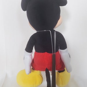 May include: A plush Mickey Mouse doll, seen from the rear, with black ears, a black body, red shorts, white gloves, and yellow shoes. The doll is mounted on a wooden stand with a metal support.