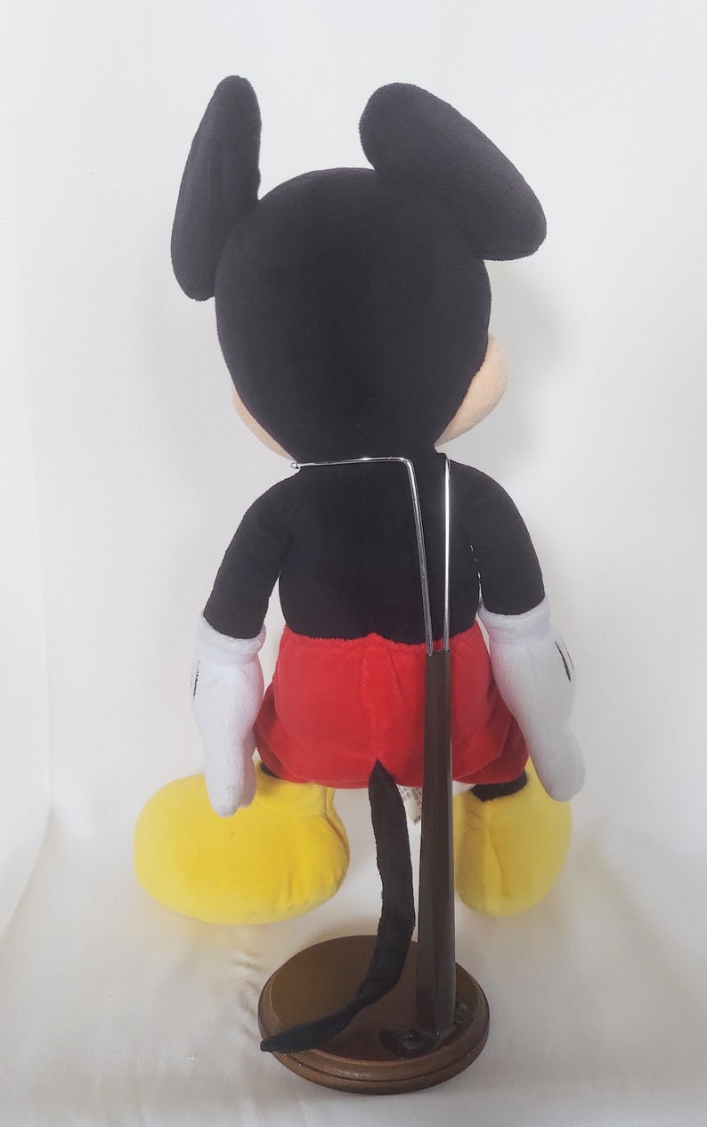 May include: A plush Mickey Mouse doll, seen from the rear, with black ears, a black body, red shorts, white gloves, and yellow shoes. The doll is mounted on a wooden stand with a metal support.