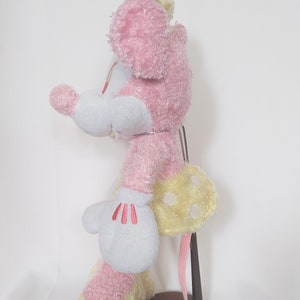 May include: A plush toy mouse in a side profile, featuring pink fur, white gloves with red accents, and a yellow polka-dotted skirt. The toy is mounted on a brown stand, showcasing its design and details.